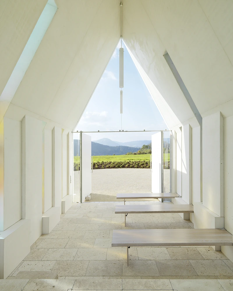 Heller Sichtbetonraum mit klarer Geometrie, schrägen Wänden und Blick in die Landschaft